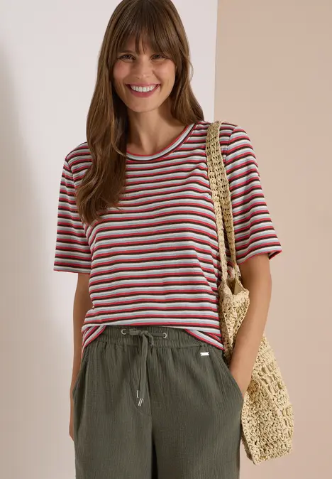 Junge Frau lächelt, trägt gestreiftes T-Shirt, olivgrüne Hose und Basttasche.