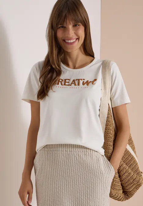 Kobieta w białej koszulce 'CREAtive', beżowej spódnicy i słomkowej torbie, uśmiechnięta.