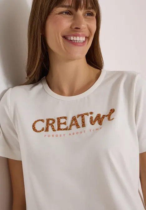 Kobieta uśmiecha się, ma na sobie biały T-shirt z napisem 'CREATIVE' z miedzianych cekinów.