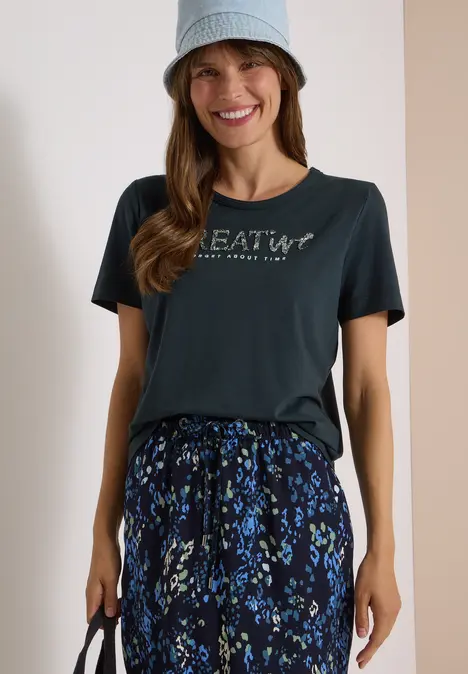 Fröhliche Frau im Bucket-Hut trägt schwarzes T-Shirt mit 'CREATIVE'-Schriftzug und gemusterte Hose.