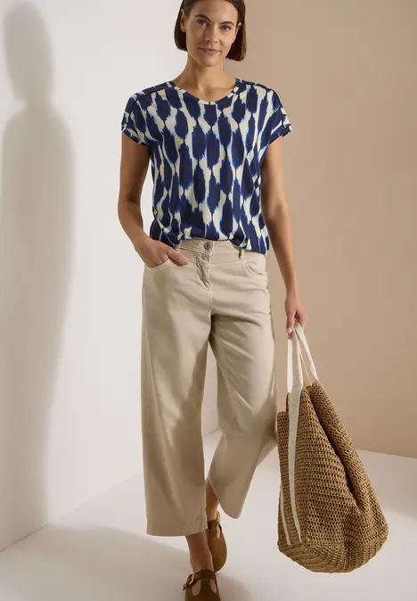 Frau trägt blau-weiß gemustertes Shirt, beige Culotte-Hose, Sandalen und großen geflochtenen Strandkorb-Tasche