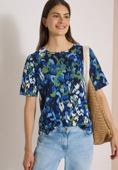Frau in geblümtem blau-grünem T-Shirt, Jeans und großer Strohtasche vor hellem Hintergrund lächelnd.