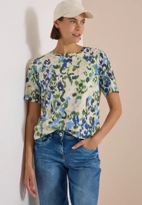 Frau mit geblümtem T-Shirt, Jeans und hellkrem Cap vor hellem Hintergrund.