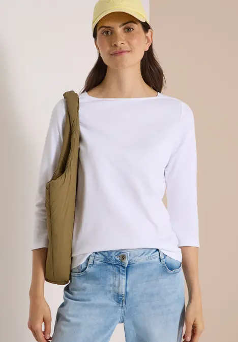 Junge Frau in Jeans, weißem 3/4-Shirt, gelber Cap und grünem Stoffbeutel vor hellem Hintergrund.