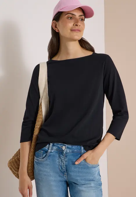 Frau trägt schwarzes 3/4-Arm-Oberteil, blaue Jeans, rosa Cap und Korbtasche vor hellem Hintergrund.