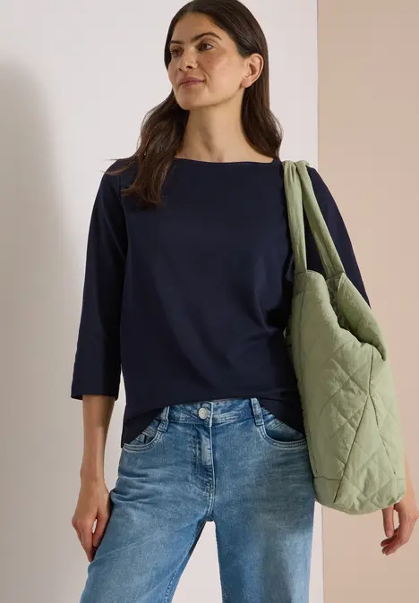 Frau trägt dunkelblaues Dreiviertelarm-Shirt, Jeans und große gesteppte grüne Tasche über Schulter.