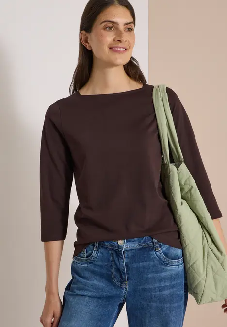 Frau mit dunkelbraunem 3/4-Arm-Shirt, blauen Jeans und grüner gesteppter Tasche lächelt.