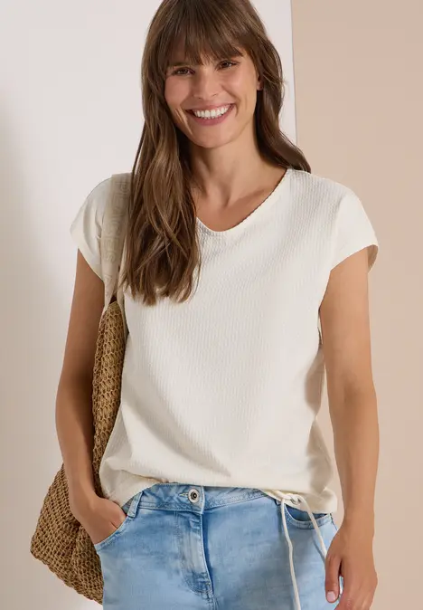 Lächelnde Frau in weißem T-Shirt und Jeans mit Strohtasche vor hellem Hintergrund.