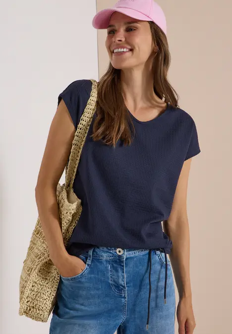 Frau in Jeans, dunkelblauem T-Shirt und rosa Cap mit Strohtasche, lächelt freudig.
