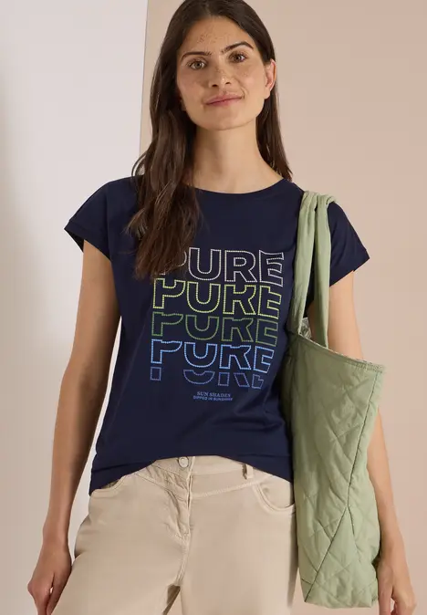Frau trägt dunkelblaues T-Shirt mit 'PURE'-Schriftzug, beige Hose und hellgrüne Stepp-Umhängetasche.