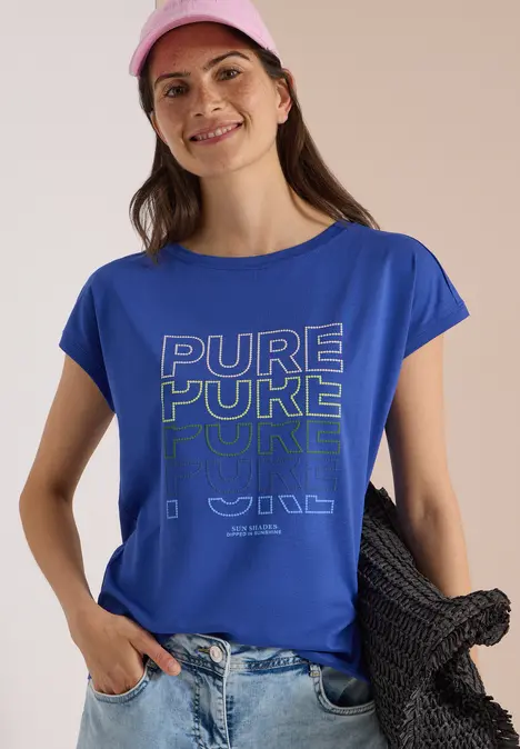 Frau in leuchtend blauem T-Shirt mit 'PURE POKE'-Print, rosa Cap, hält schwarze Strickjacke.