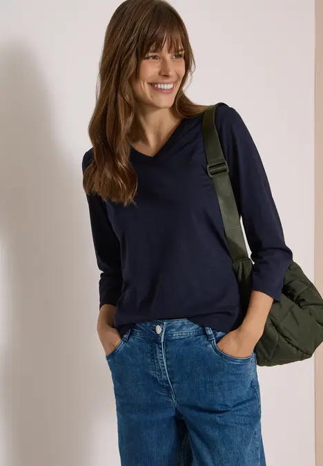Frau mit dunkelblauem Pullover und Jeans, lächelt entspannt mit grünem Rucksack über der Schulter.