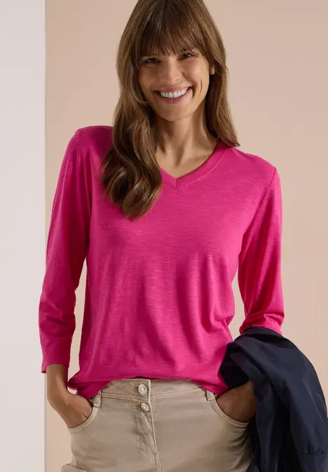 Lächelnde Frau in pinkem Langarmshirt mit V-Ausschnitt, beigen Hosen und dunklem Blazer über dem Arm.