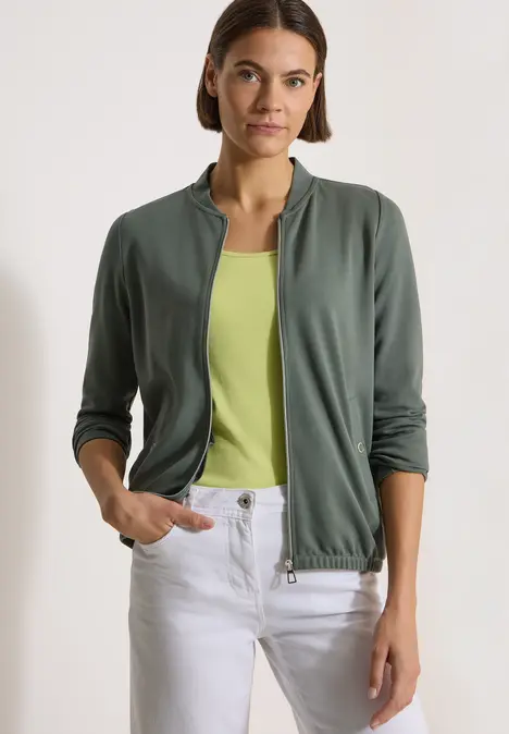 Frau trägt khakigrüne Jacke über hellgrünem Top und weiße Hose, Hände in den Taschen.