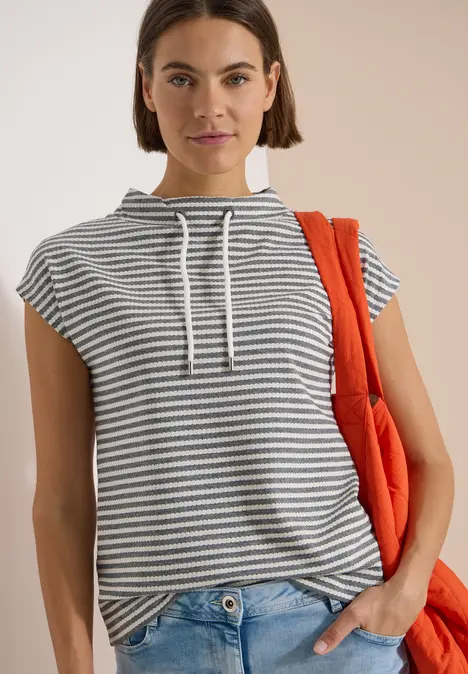 Person trägt gestreiftes Ärmelloses Kapuzenshirt, Jeans, hält eine orangene Tasche, vor beigem Hintergrund.