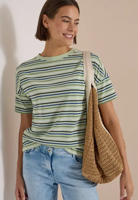 Frau in gestreiftem T-Shirt und Jeans trägt eine geflochtene Tasche und lächelt.