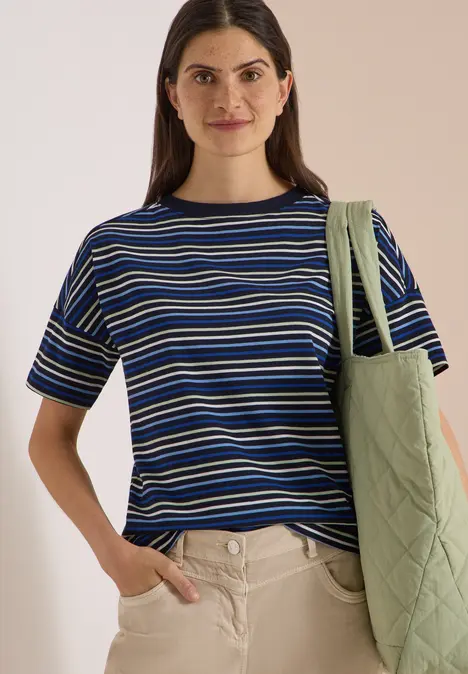 Frau im blau-weiß gestreiften T-Shirt mit grüner Stepp-Tasche über der Schulter, lächelnd.