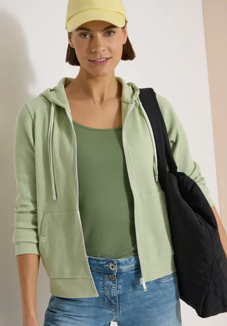 Person vor weißem Hintergrund mit beiger Mütze, hellgrüner Kapuzenjacke, grünem Top, Jeans und schwarzem Beutel.