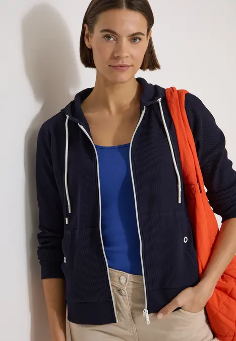 Person trägt dunkelblauen Hoodie, blaues Top und helle Hose, hält rote Tasche über der Schulter.