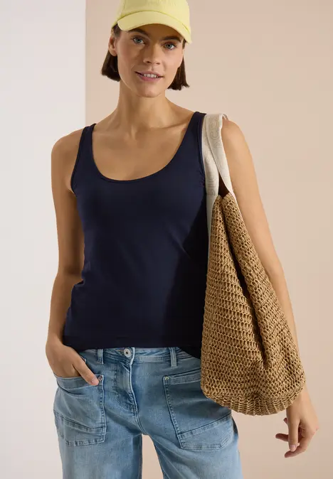 Frau im schwarzen Tanktop, Jeans und Strohtasche über der Schulter.