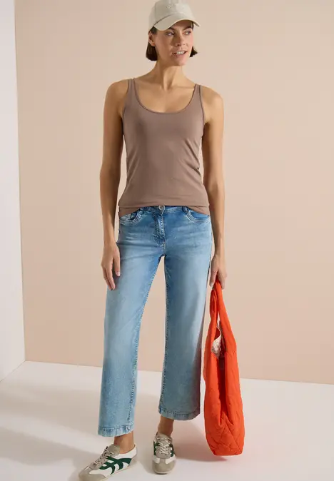 Eine Person in braunem Top, blauen Jeans und Sneakers hält eine rote Tasche vor hellem Hintergrund.