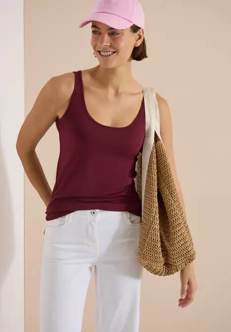 Frau mit burgunderfarbenem Top, weißer Hose, pinkem Cap und grober Strohtasche.