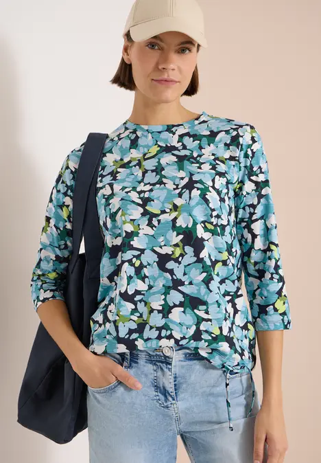 Frau mit blauer floral bedruckter Bluse, Jeans und Shopper-Tasche vor hellem Hintergrund.