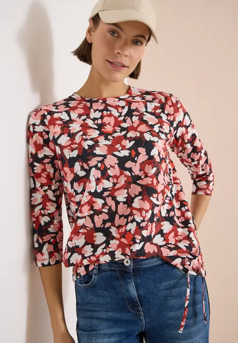 Frau in Blütenmuster-Bluse mit 3/4-Ärmeln und Jeans vor hellem Hintergrund.