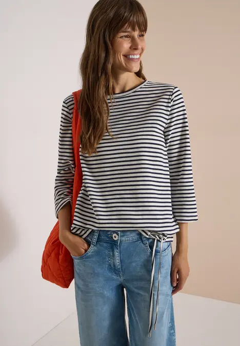 Frau in gestreiftem Longsleeve, Jeans und roter Tasche, die lächelnd in die Kamera schaut.