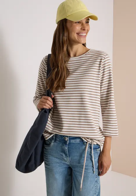 Junge Frau mit beiger-weißer Ringelbluse, blauen Jeans, gelber Cap und dunkelblauer Tasche lächelnd vor hellem Hintergrund.