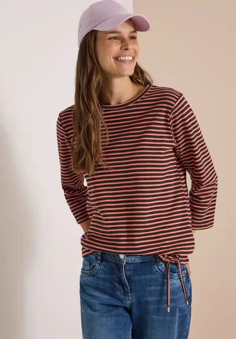 Junge Frau mit gestreiftem Langarmshirt, Jeans und lila Baseballcap, lächelnd.