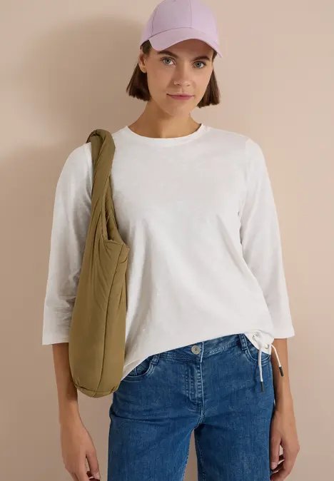Junge Frau in weißem Langarmshirt, blauen Jeans, lila Kappe und grünem Beutel vor beigem Hintergrund.