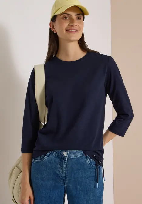 Eine lächelnde Frau trägt eine marineblaue Langarmbluse, blaue Jeans, eine gelbe Cap und eine Umhängetasche.