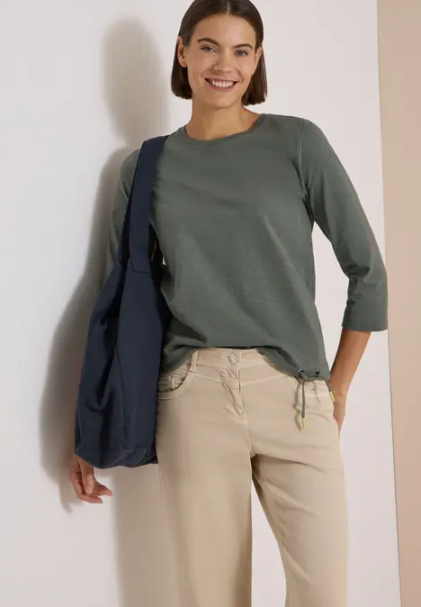 Lächelnde Frau in grünem 3/4-Arm-Shirt, beiger Hose und blauer Umhängetasche.