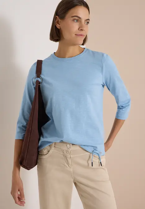 Frau im hellblauen 3/4-Arm-Shirt, beige Hose und brauner Umhängetasche, schaut seitlich vor hellem Hintergrund.