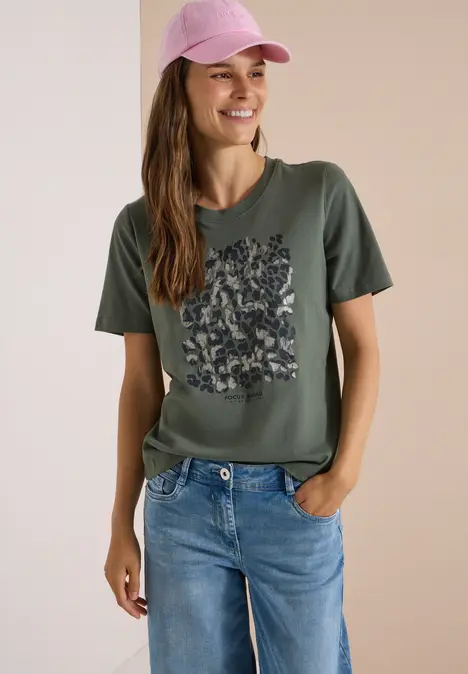 Frau mit rosa Cap, olivgrünem T-Shirt mit Blumenmuster und blauen Jeans lächelnd.