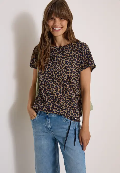 Vrolijke vrouw met lang haar, draagt een luipaardshirt en blauwe jeans tegen een lichte achtergrond.