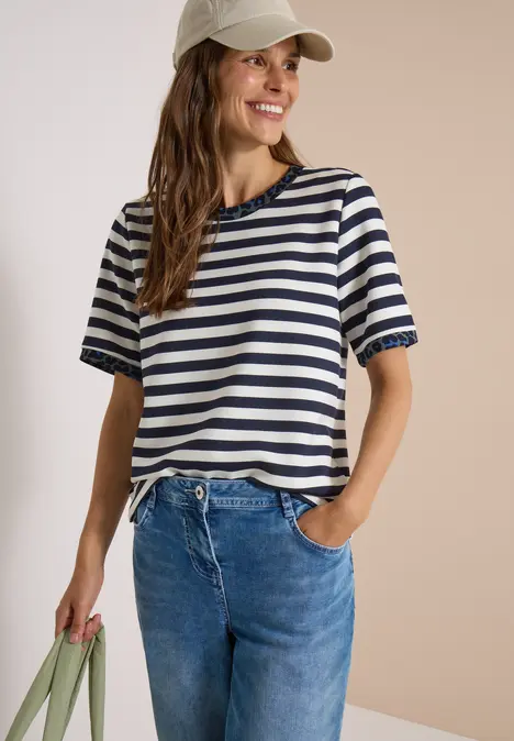 Junge Frau in gestreiftem T-Shirt, Jeans und Baseballcap, lächelnd mit grüner Tasche.