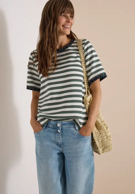 Frau in gestreiftem T-Shirt, blauen Jeans und Strohtasche lächelt seitlich.