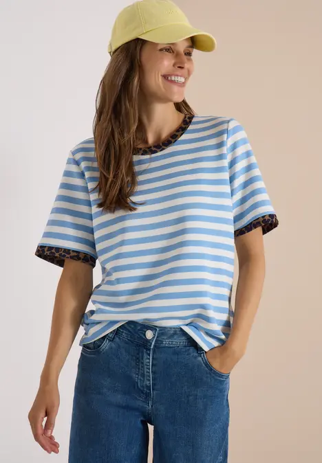 Frau mit gelber Cap, blau-weiß gestreiftem T-Shirt mit Leoprint-Bündchen und Jeans lächelnd posierend.