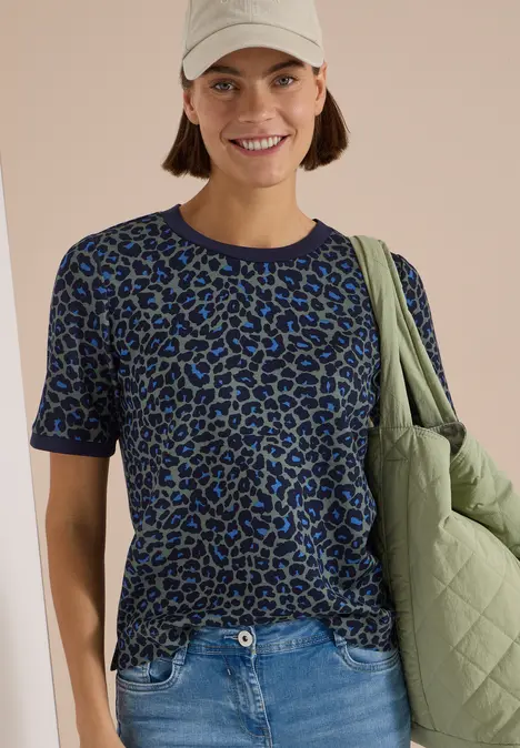 Frau lächelt in beiger Mütze, blau-grünem Leo-Shirt, Jeans und grünem Steppbeutel.