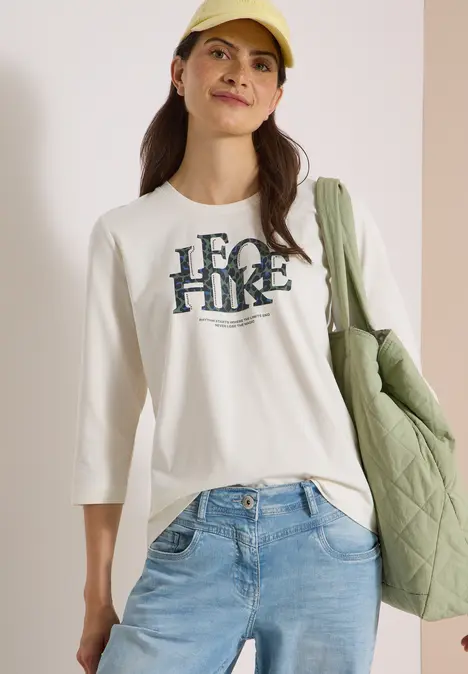 Frau in Jeans, weißem 3/4-Shirt mit 'HERO HIKE'-Schriftzug, gelber Mütze und grüner, gesteppter Tasche.
