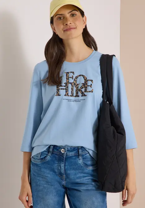 Frau mit beiger Kappe, hellblauem 3/4-Shirt mit Leo-Print-Schriftzug, Jeans und schwarzer Tasche vor neutralem Hintergrund.