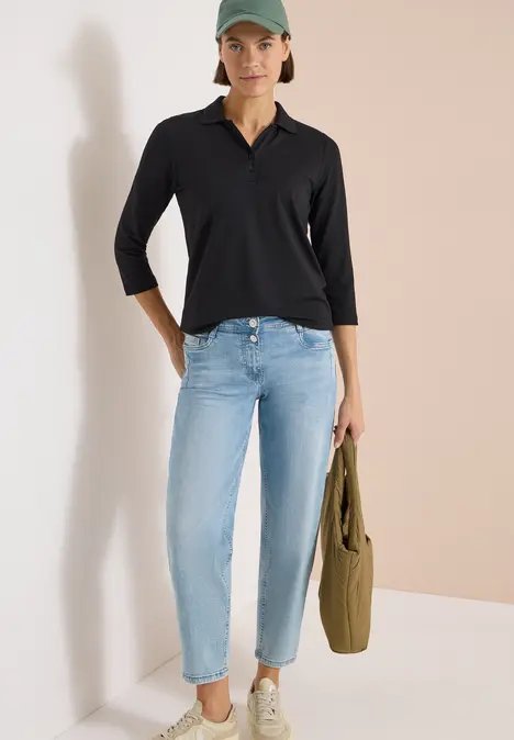 Person steht an Wand, trägt schwarzes Dreiviertelärmel-Shirt, hellblaue Jeans und hält einen olivfarbenen Stoffbeutel.