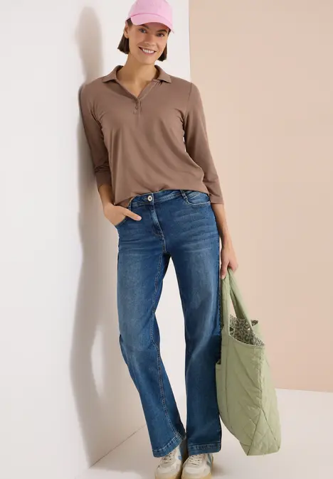 Person lehnt an Wand, trägt braunes Poloshirt, blaue Jeans und hält eine große grüne Tasche.