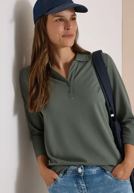 Frau lehnt an der Wand, trägt olivgrünes Poloshirt mit 3/4-Ärmeln, Jeans, blaue Cap und Schultertasche.