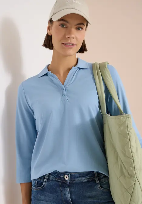 Eine Person trägt ein hellblaues Langarmshirt, Jeans und eine grüne, gesteppte Tragetasche.