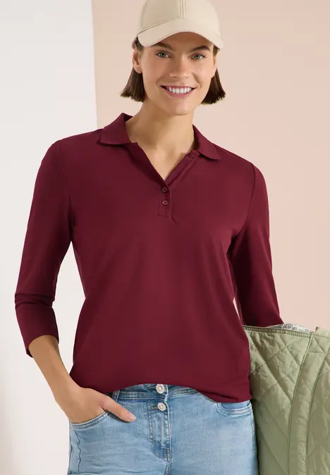 Dame trägt burgunderrote 3/4-Arm-Polobluse mit V-Knopfleiste und hellblaue Jeans, Hand in Tasche.
