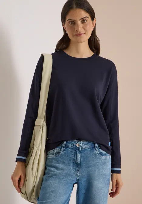 Frau in dunkelblauem Pullover und Jeans mit Beuteltasche vor neutralem Hintergrund.
