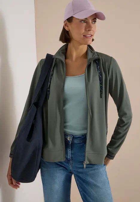 Frau mit lila Cap, grüner Zip-Jacke, hellblauem Top, Jeans und blauer Tasche.